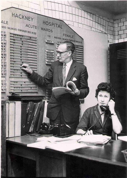 Photo:Mr L E Head, medical records officer, checking the number of available beds in the hospital on a chart specially designed by the hospital staff. Mrs Edna Gore, admissions clerk, is on the telephone seated beside him, Apr 1962 (Catalogue reference: SBHH-PG-116)